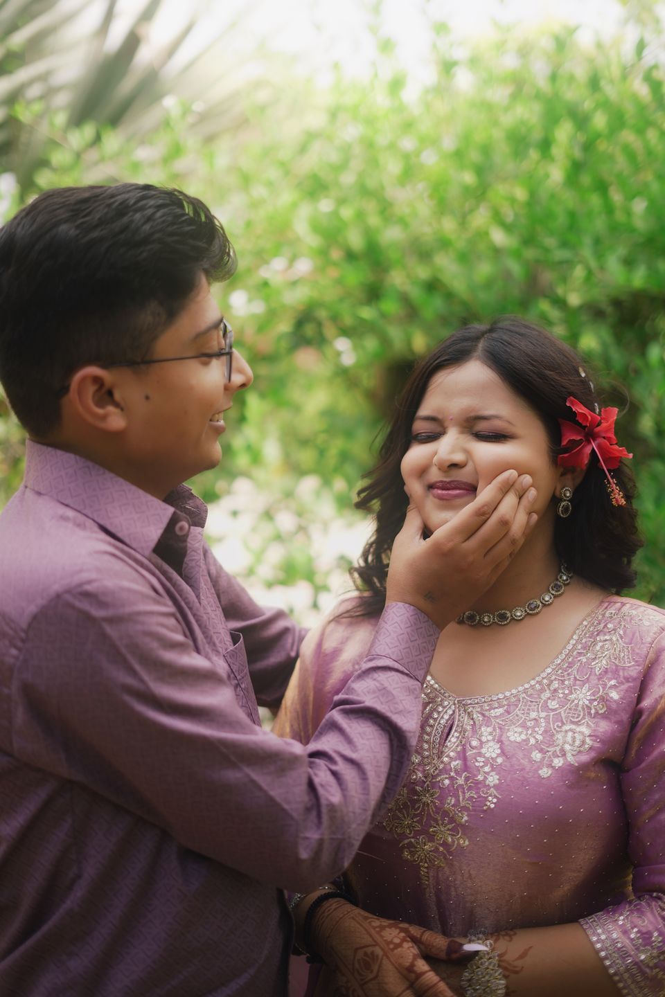 Groom gently holding the bride's face as she laughs, a red flower in her hair